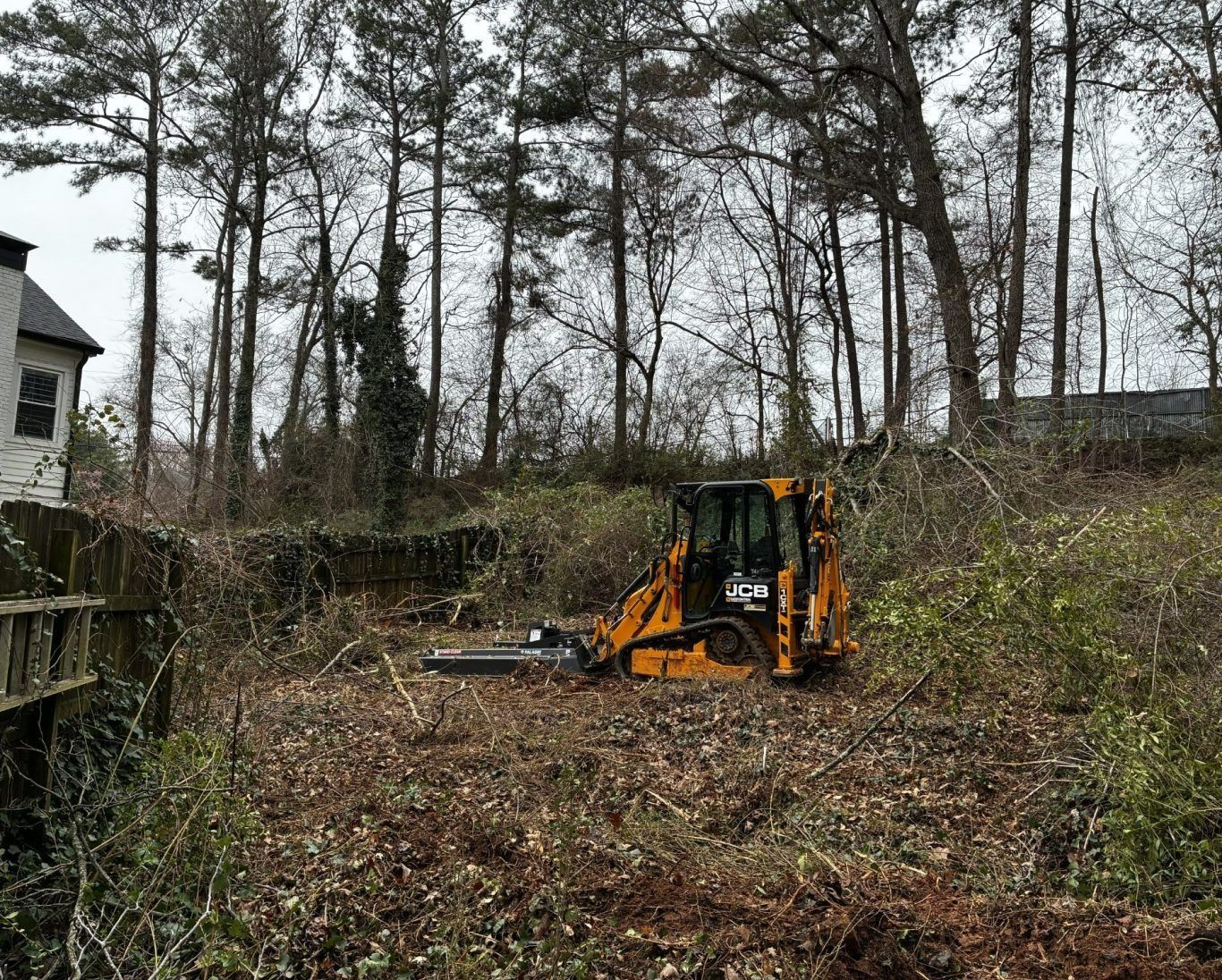 Yellow excavator clearing brush in a wooded, overgrown area near a house