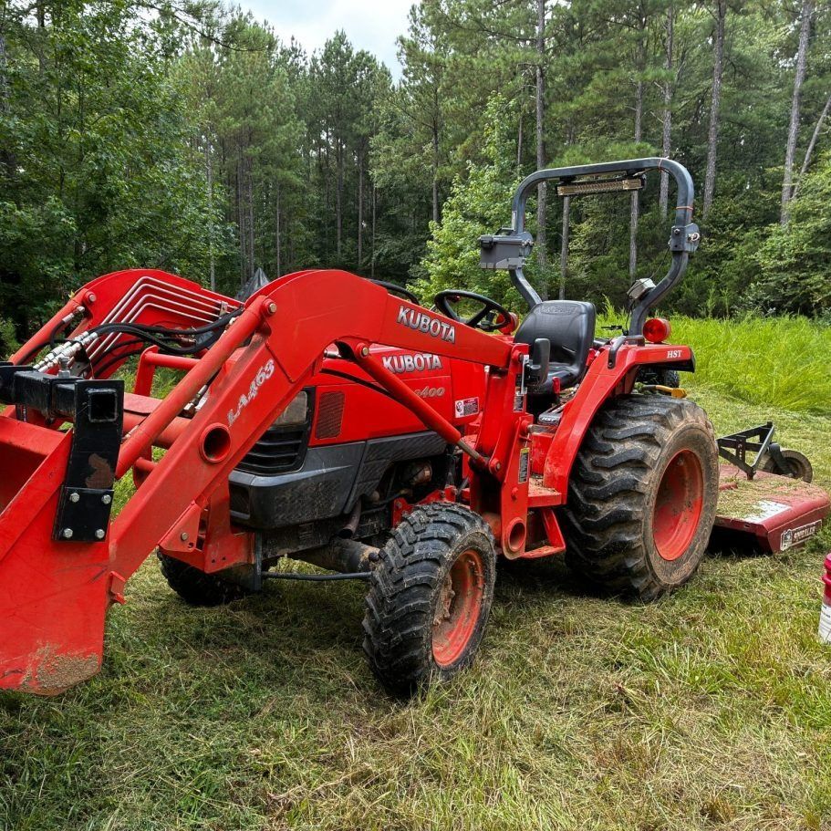 Red tractor with front loader and mower attachment parked in a grassy wooded area