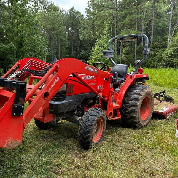 Red tractor with front loader and mower attachment parked in a grassy wooded area