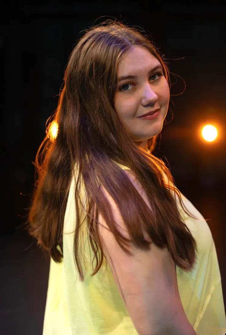 Kaitlyn Ray in a yellow top turns back and smiles under warm stage lights.