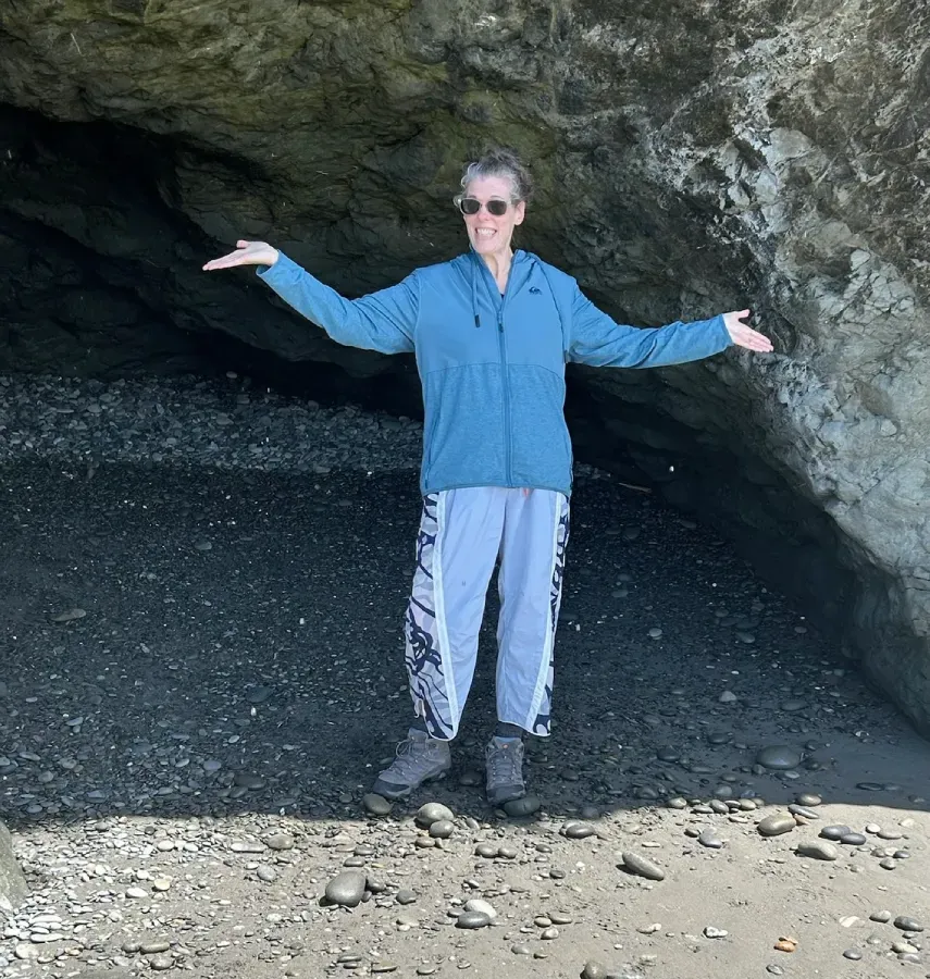 Nicole standing in a cave entrance with arms outstretched, wearing a blue jacket and gray pants.