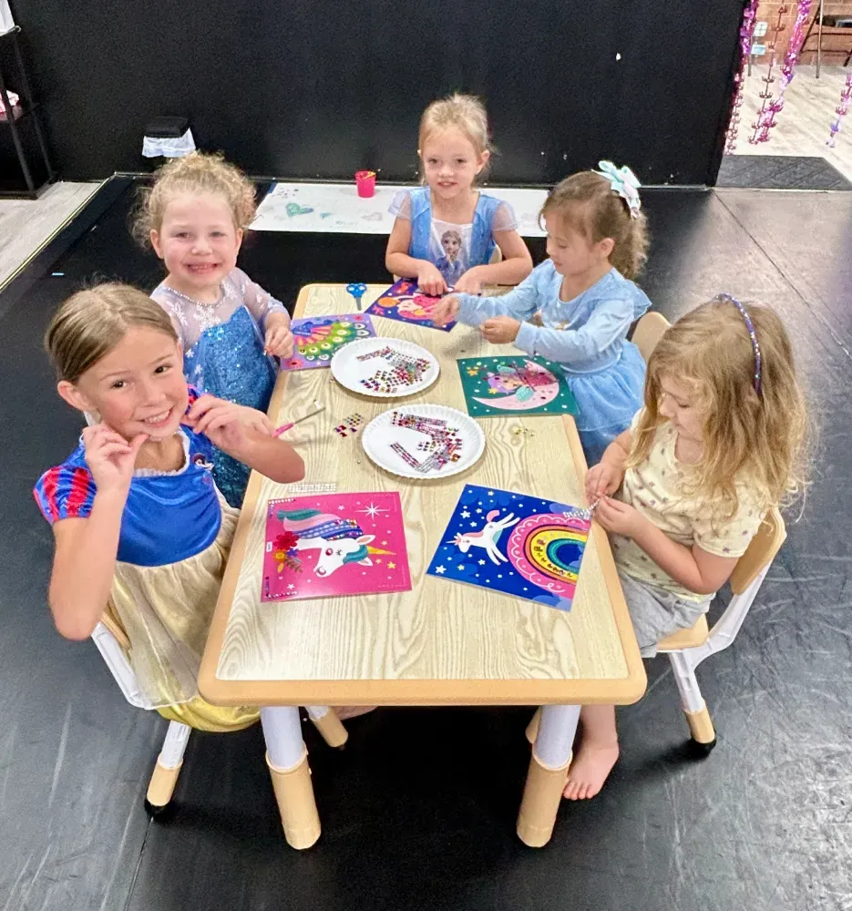 Five children sit around a small table, smiling and painting colorful crafts in a dance classroom