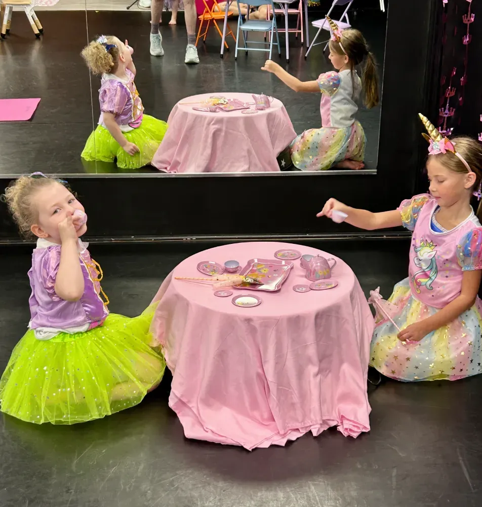 Children in pastel costumes play at a pink tea party table with a mirror reflection behind them.