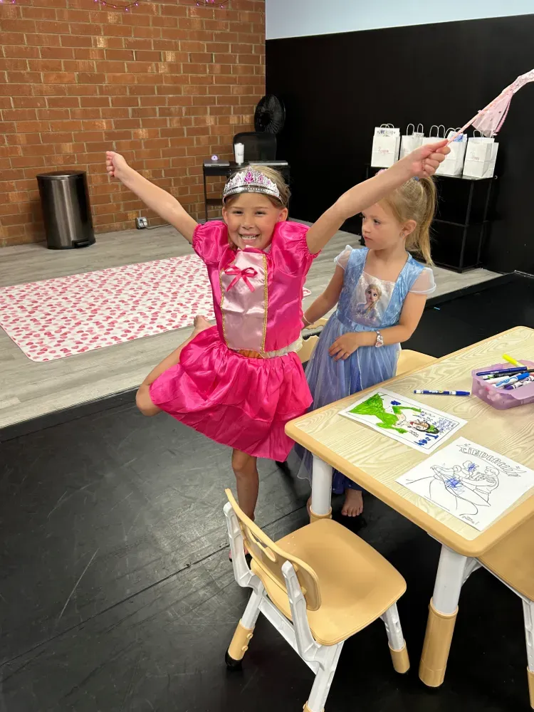 Two children in princess costumes pose with arms raised at a table in a classroom or party room.