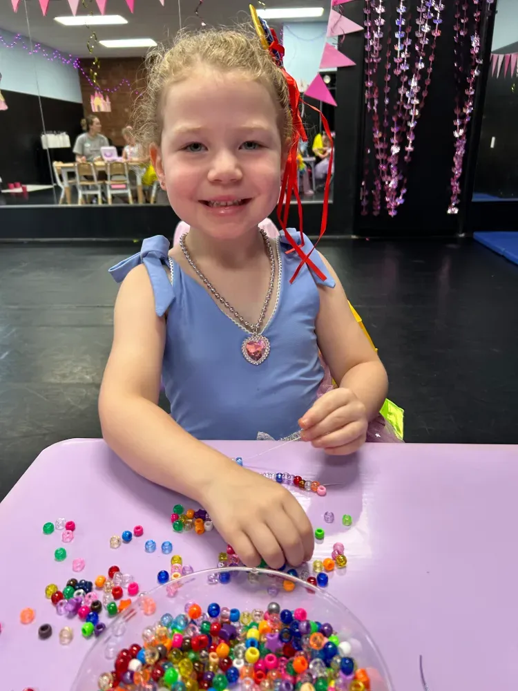 Smiling child at a table, playing with colorful beads in a bowl and scattered across a purple surface.