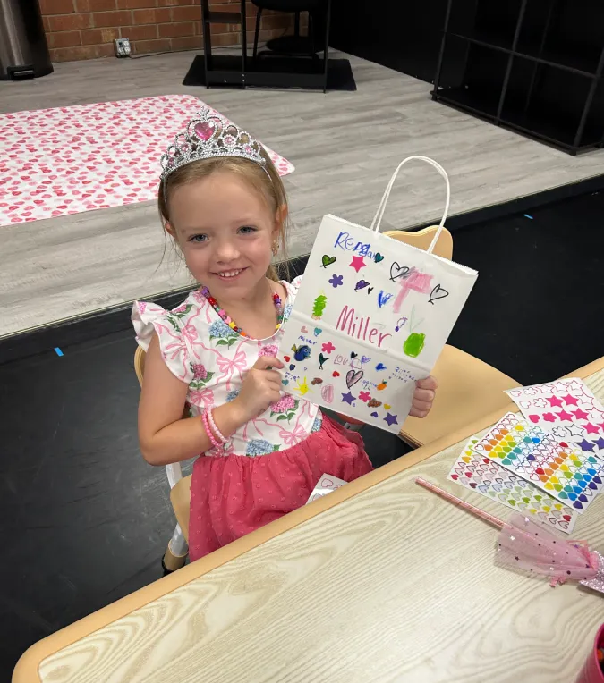 Smiling child holding a decorated gift bag at a table with colorful craft supplies
