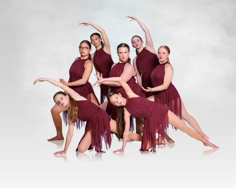 Six dancers in burgundy costumes pose in a synchronized contemporary dance formation on a white background