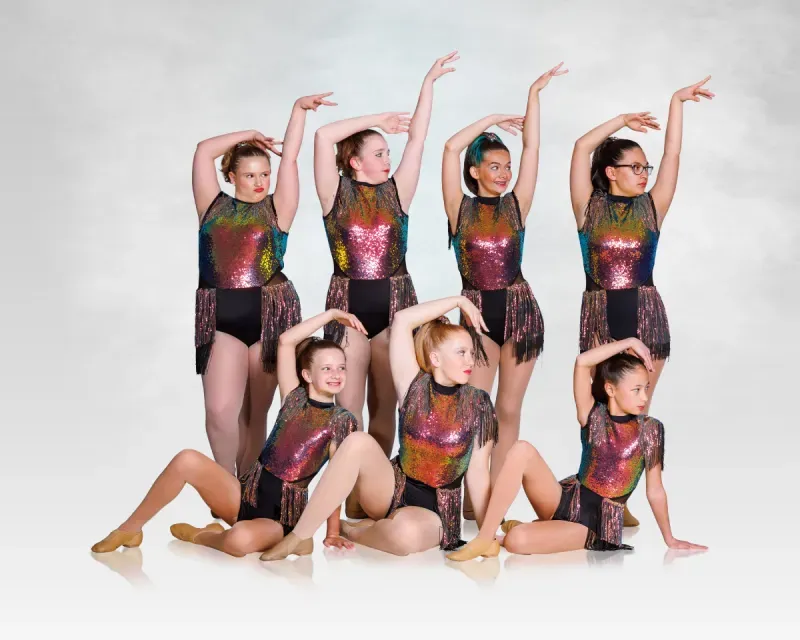 Seven dancers in glittery leotards pose in a studio, arms raised in synchronized formation.