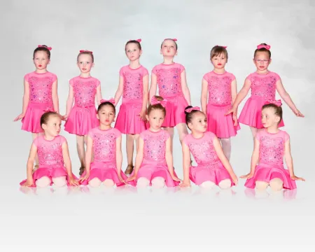 A group of children in matching pink dance costumes posed in two rows on a white background