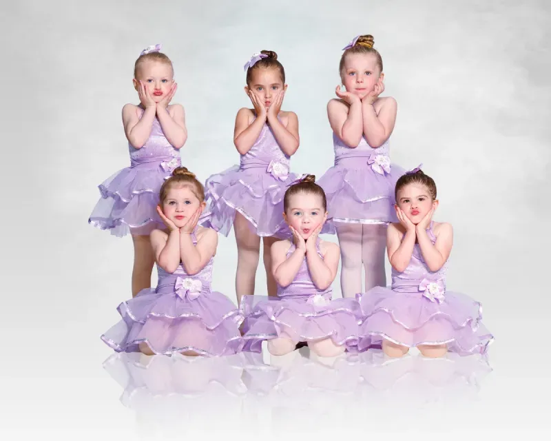 Six young dancers in lavender tutus pose on a white background, hands by their cheeks.