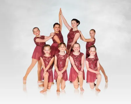 Seven dancers in burgundy costumes posing against a white backdrop, one raised in a high center pose.