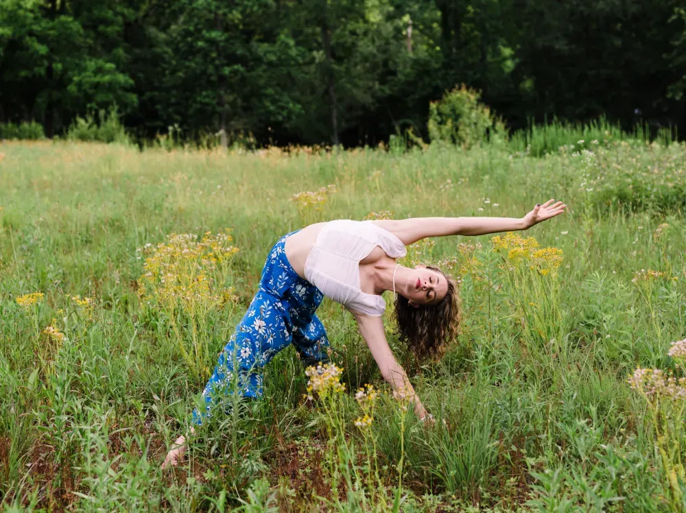 Sidney bending sideways in a grassy field, wearing blue patterned pants and a white top, with arms extended.
