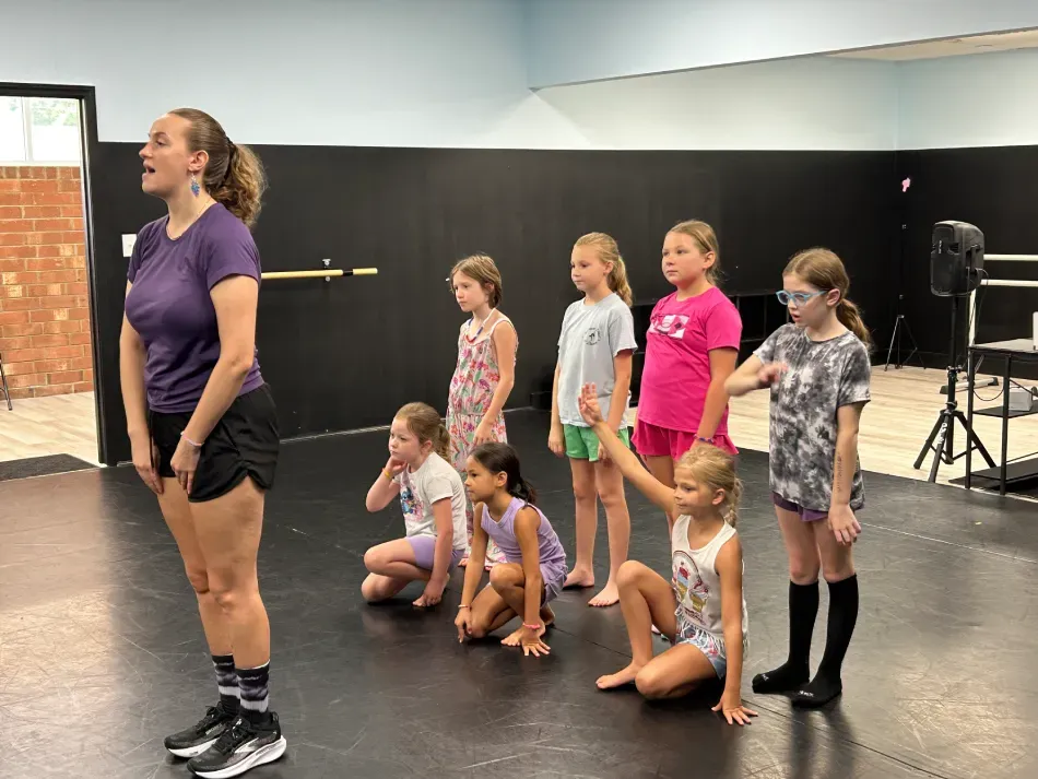 A dance instructor leading a group of children in a dance studio through a routine