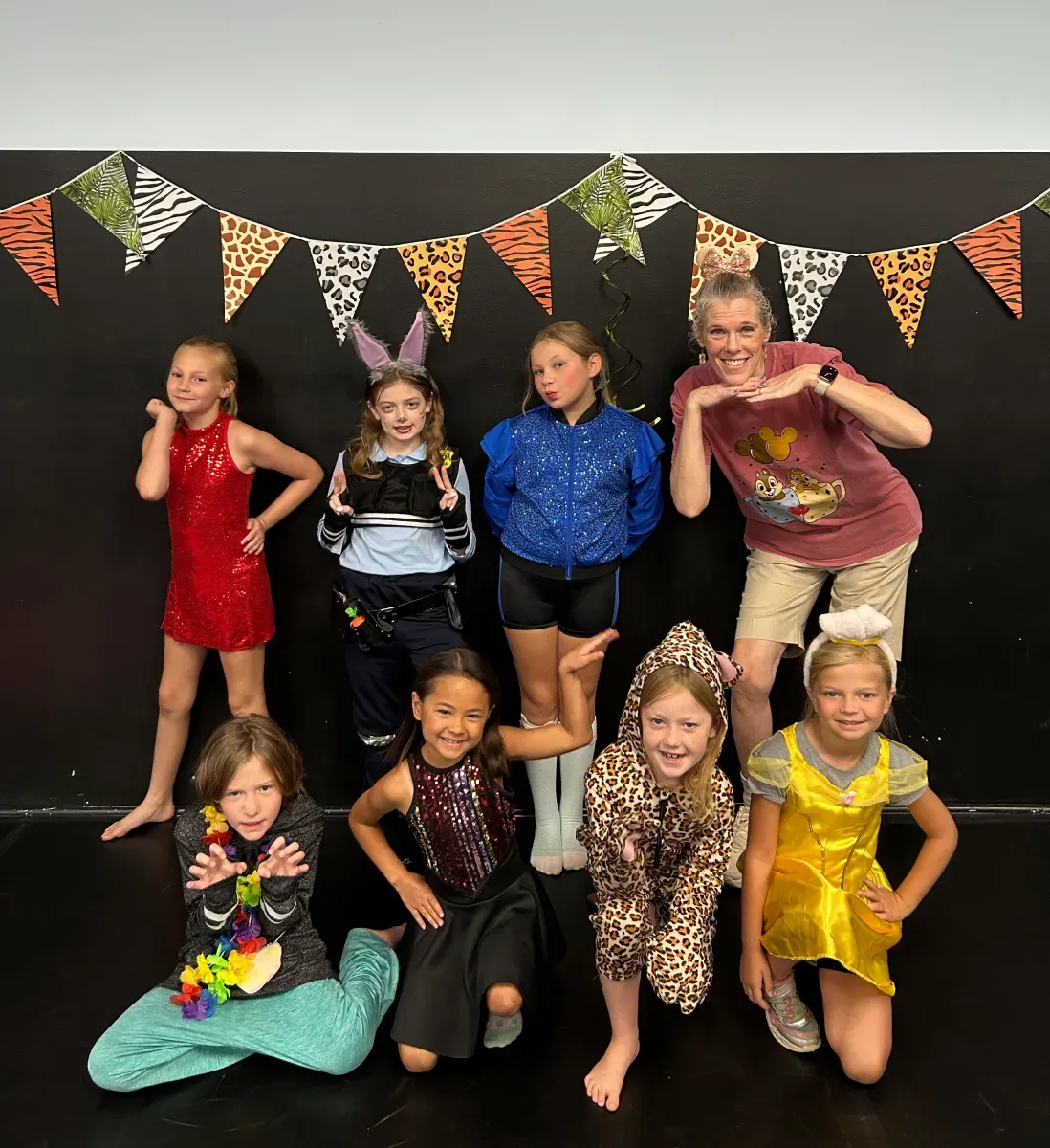 Seven children posing in colorful animal themed costumes in front of a black backdrop with animal print bunting