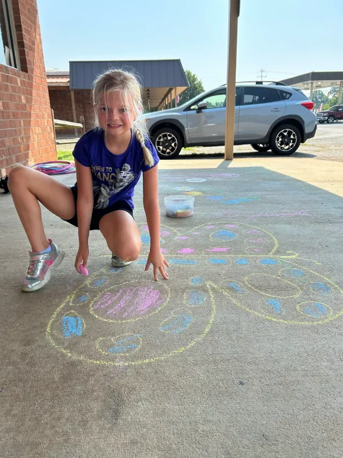 Girl crouching on a driveway beside colorful chalk drawings with a parked car in the background