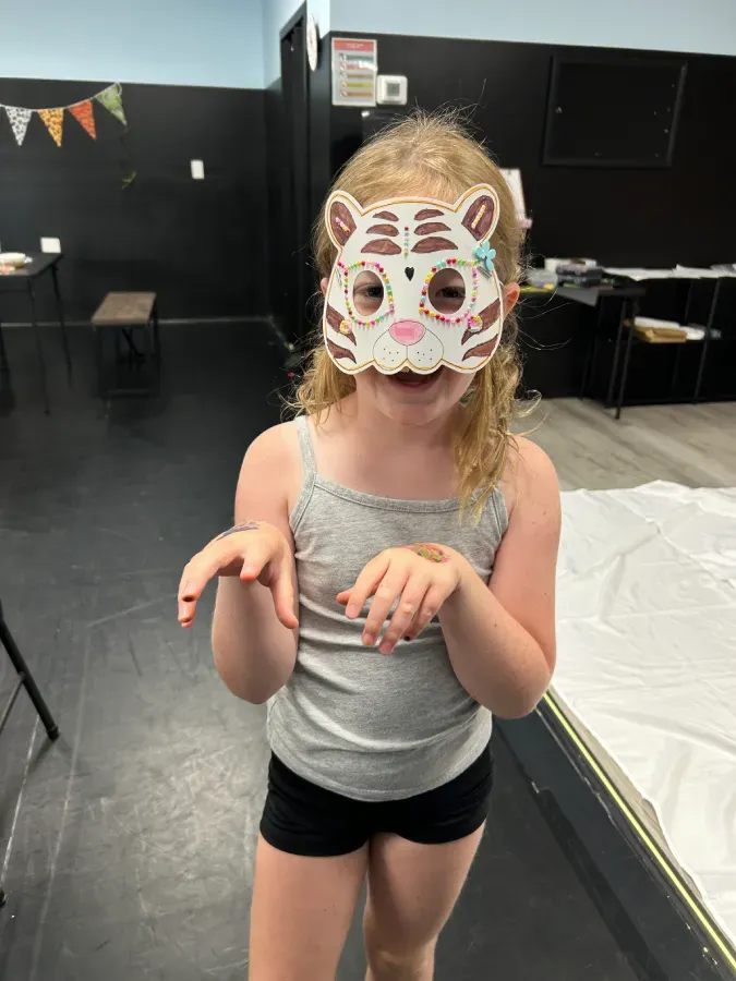 Child wearing a handmade cat mask and gray tank top, posing indoors on a stage with a playful hand gesture