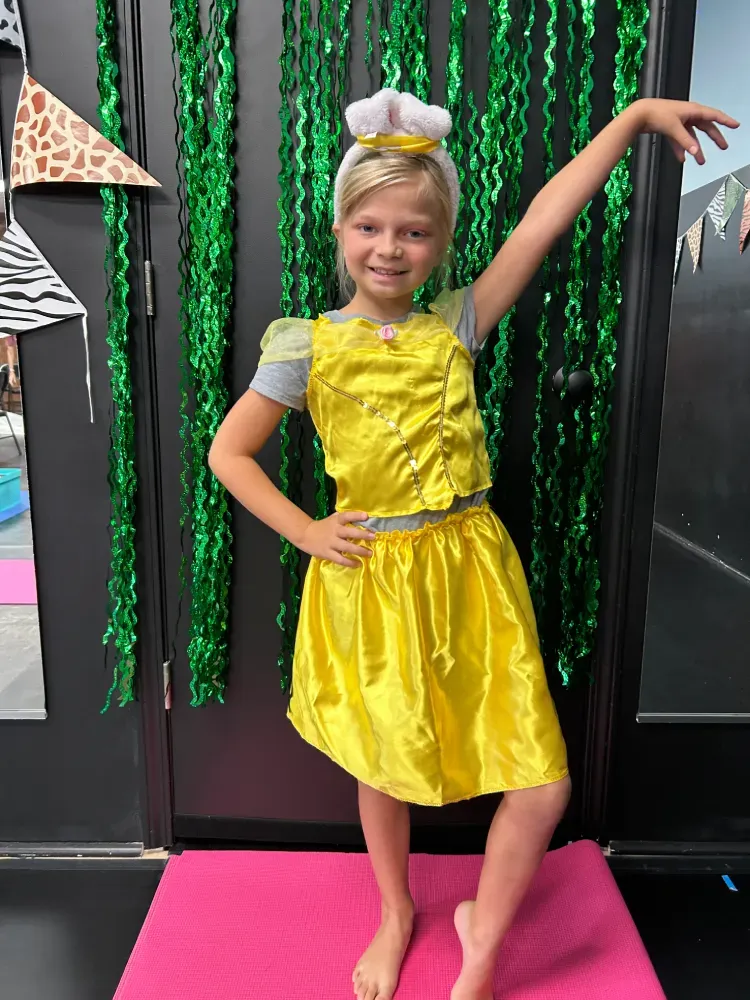 Child in a yellow dress and silver headpiece posing on a pink mat with green streamers behind.