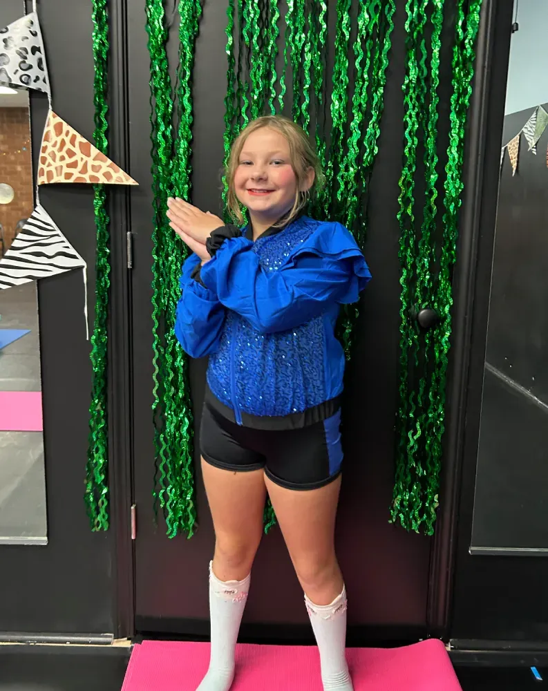 A child in blue jacket posing with arms crossed in front of green tinsel backdrop