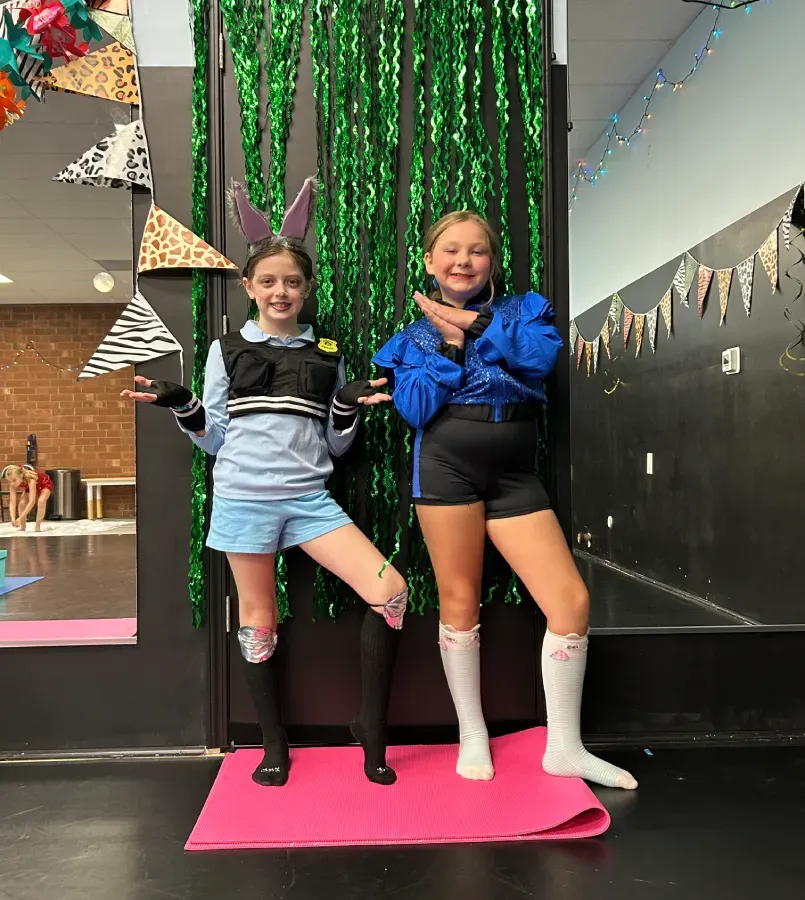 Two dancers pose on a pink mat in a decorated studio, wearing black and blue outfits.