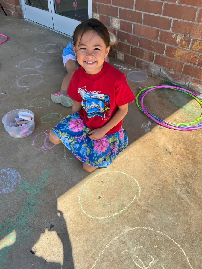 Smiling child in a red shirt sitting outside on pavement with chalk drawings and colorful hula hoops nearby