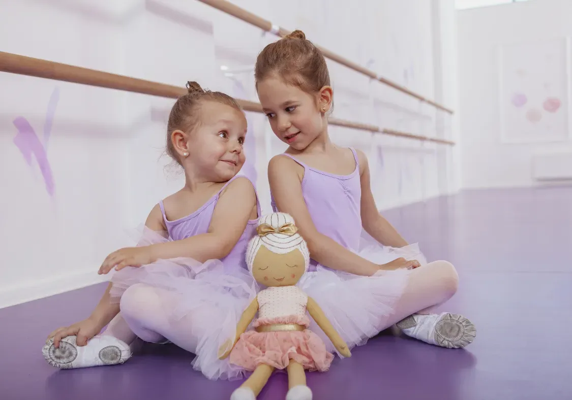 Two ballerinas in lavender tutus sit on a purple studio floor, smiling at each other beside a doll.