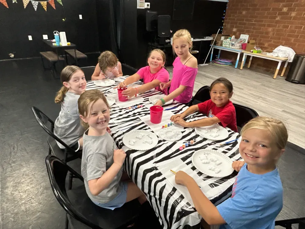 Children seated at a table with zebra-print cloth, smiling and painting in a dance studio classroom