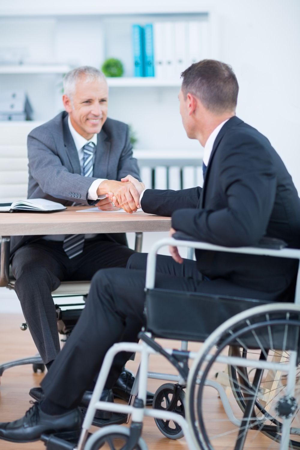 Two men in business attire shake hands across a desk, one seated in a wheelchair, in a bright office.