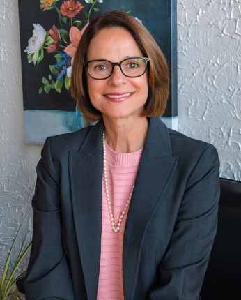 Woman in glasses and dark blazer standing before a floral painting and white textured wall.