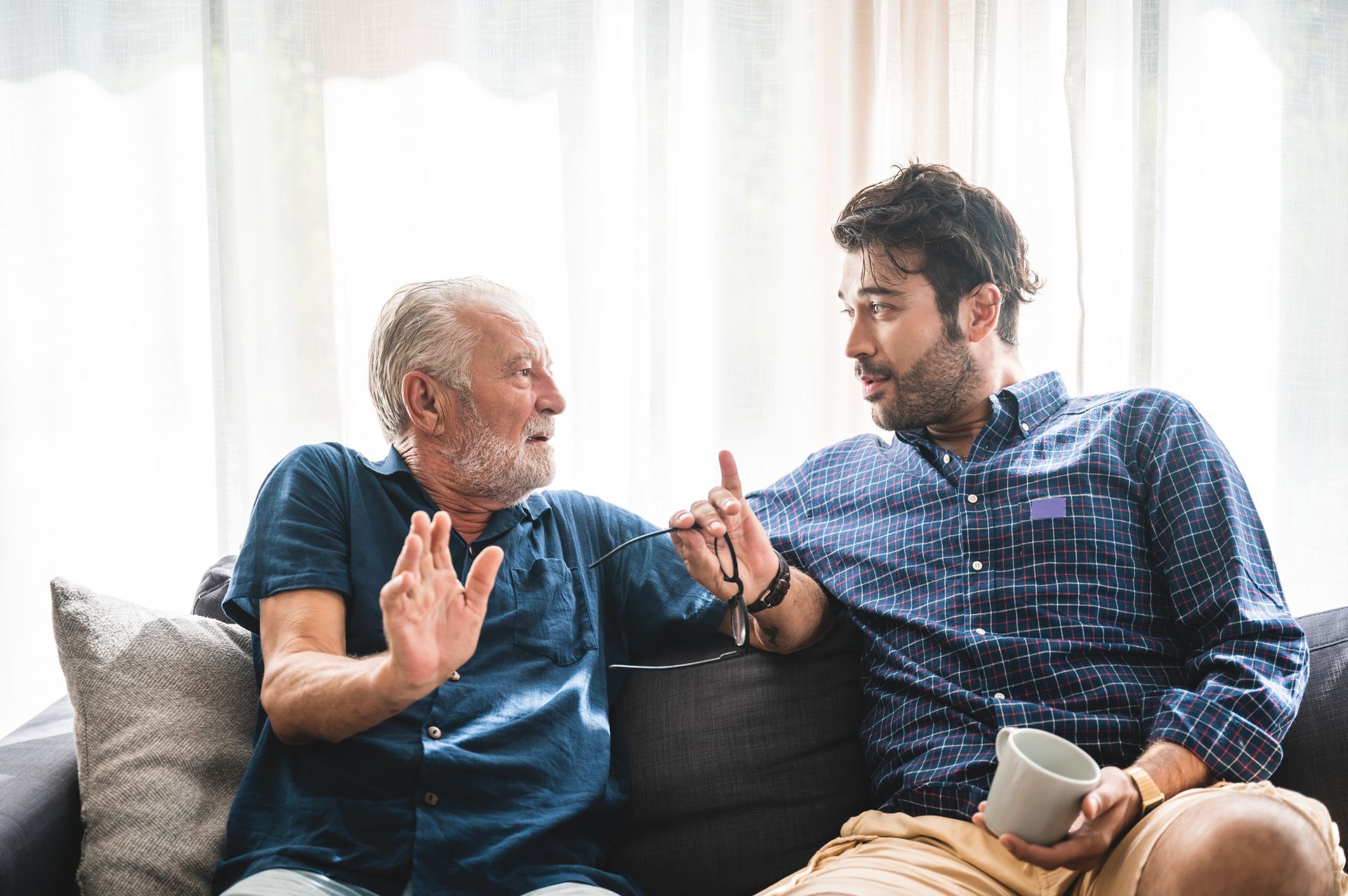 Two men talking on a couch, gesturing with their hands in a bright living room.