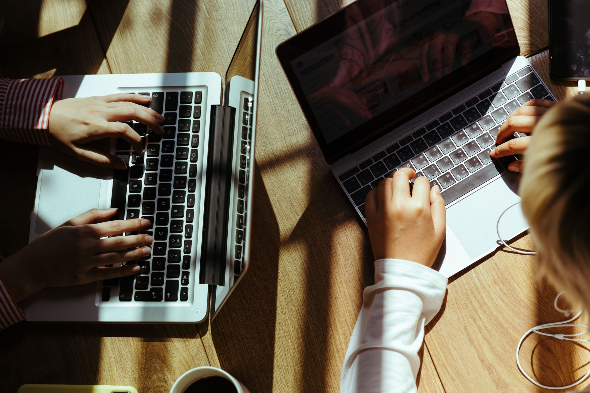 Two people typing on laptops at a sunlit wooden table, with coffee cups and cables nearby