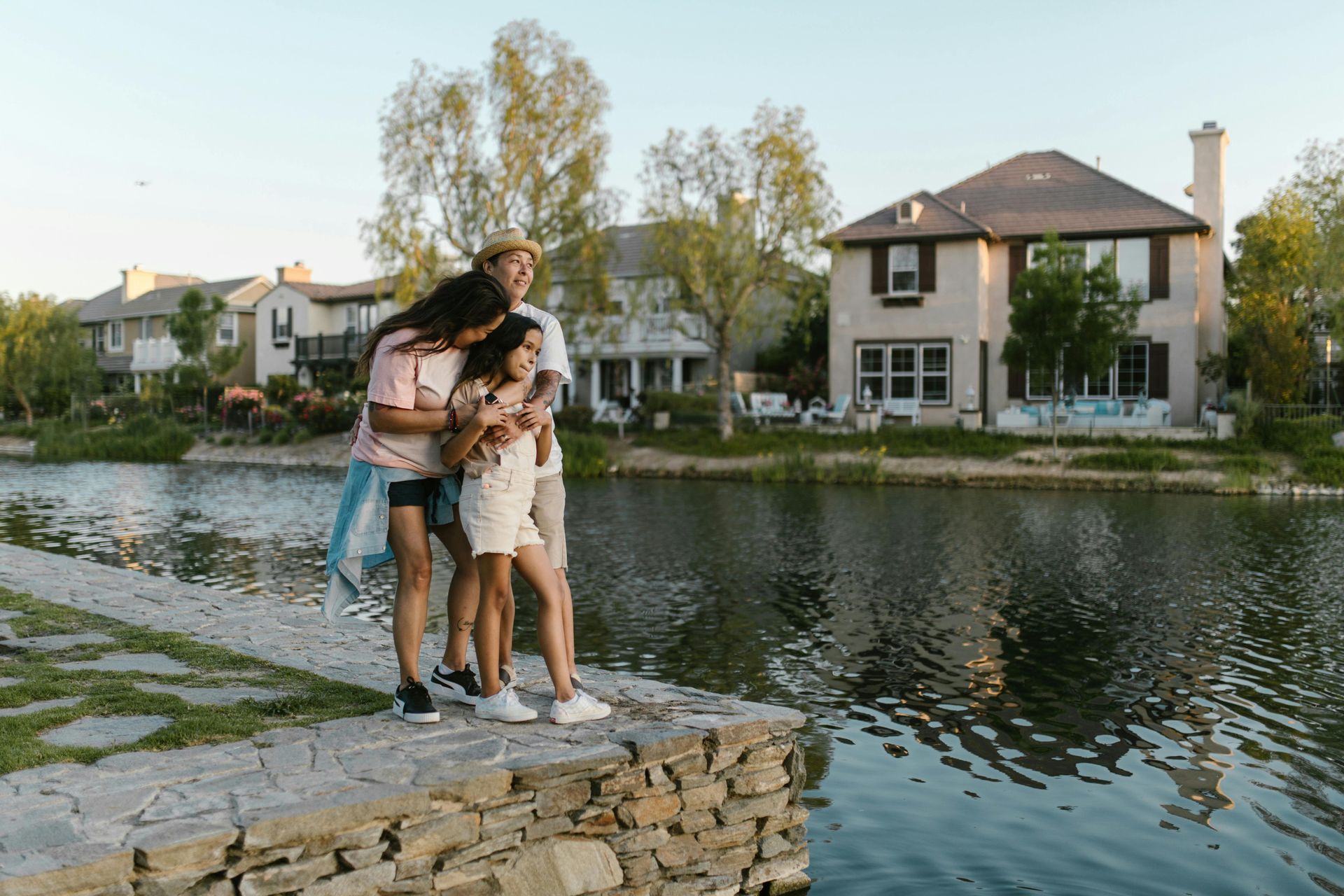 Couple embracing on a stone ledge by a calm river with houses and trees in the background
