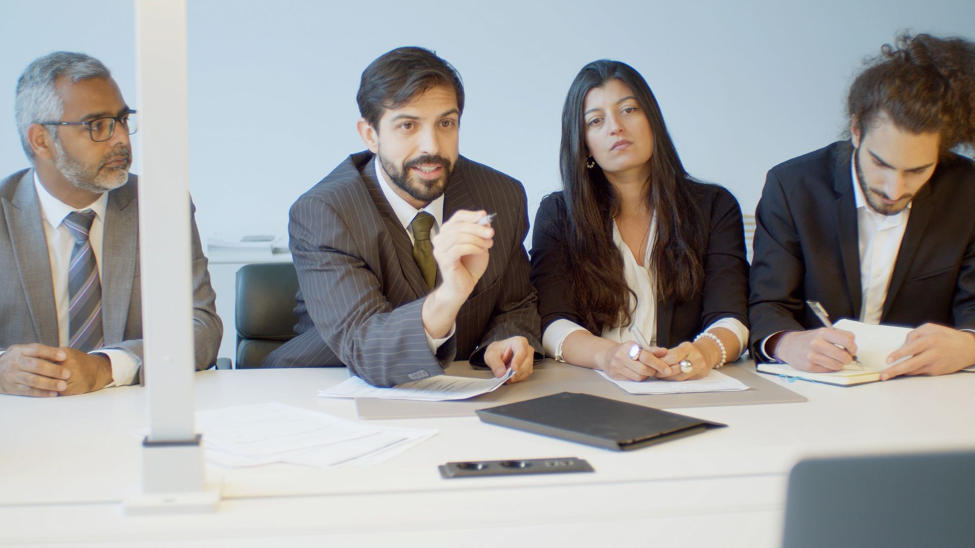 Four colleagues in business attire discuss documents around a conference table in a bright office.