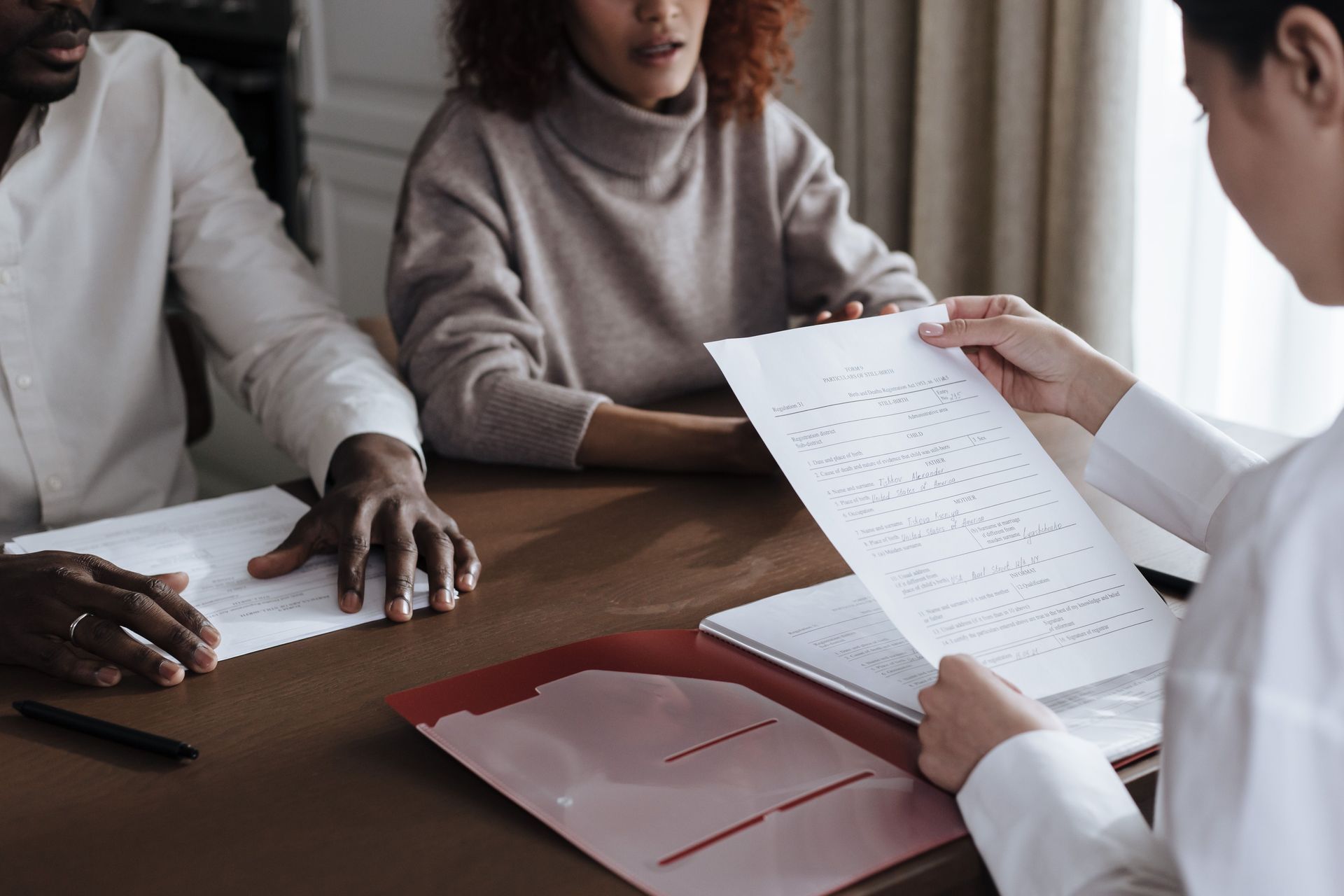 Three people review documents at a wooden table in a bright meeting room.