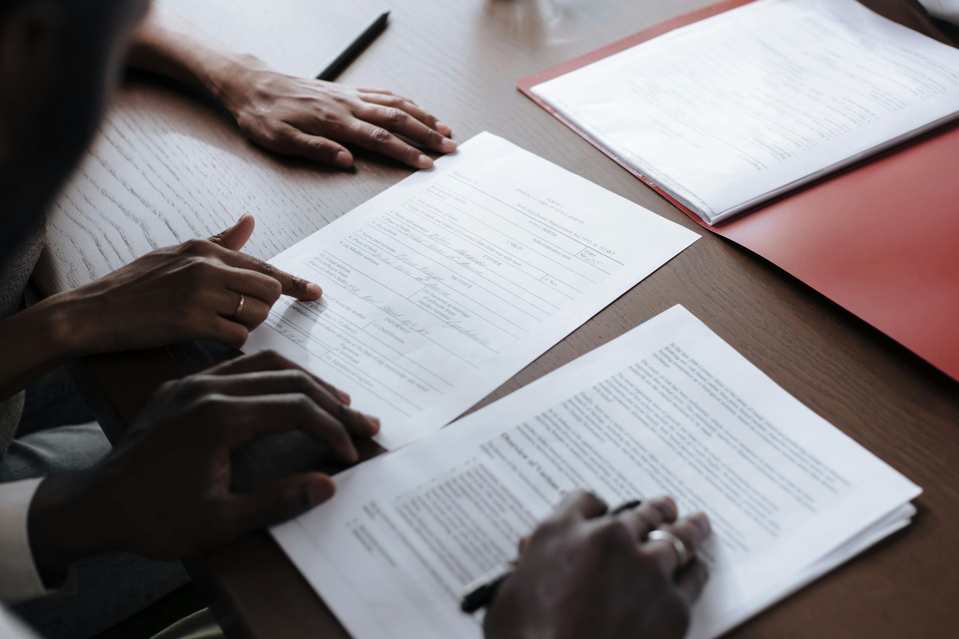Hands reviewing paperwork on a desk with printed documents and a red notebook