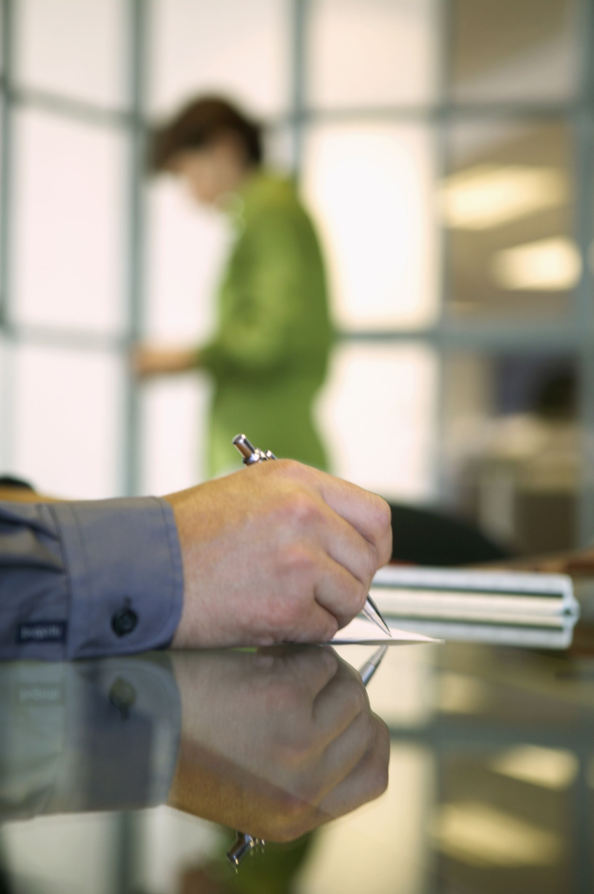 Hand signing a document at a reflective desk, with a blurred person in green standing in the background