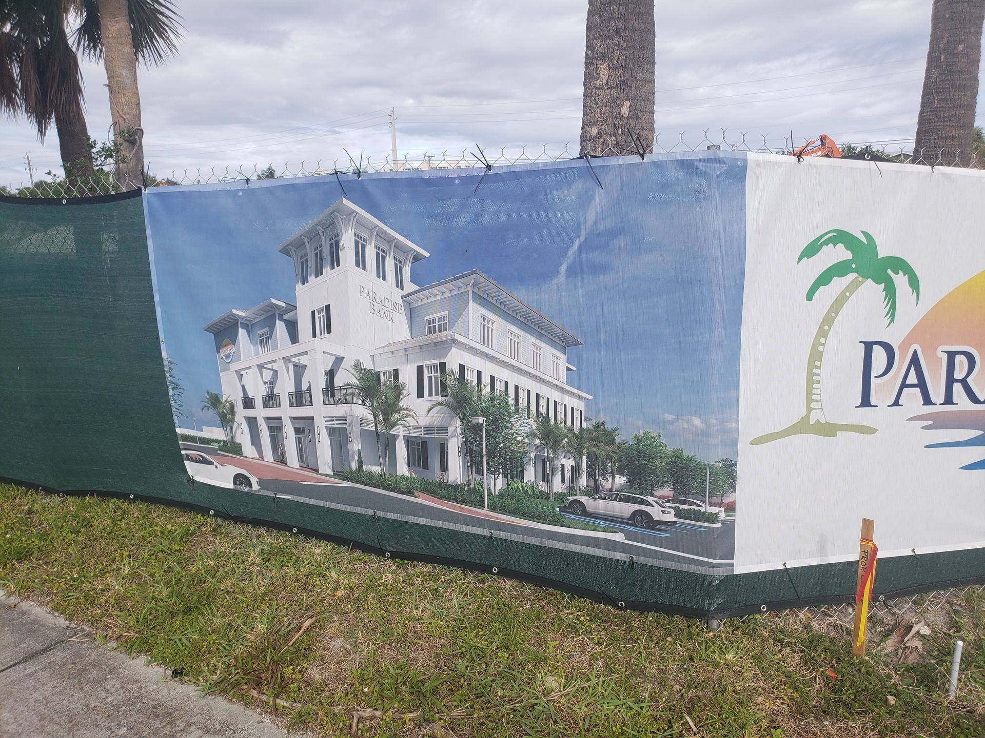 A construction site fence with renderings of a white building and a logo with a palm tree and sun.