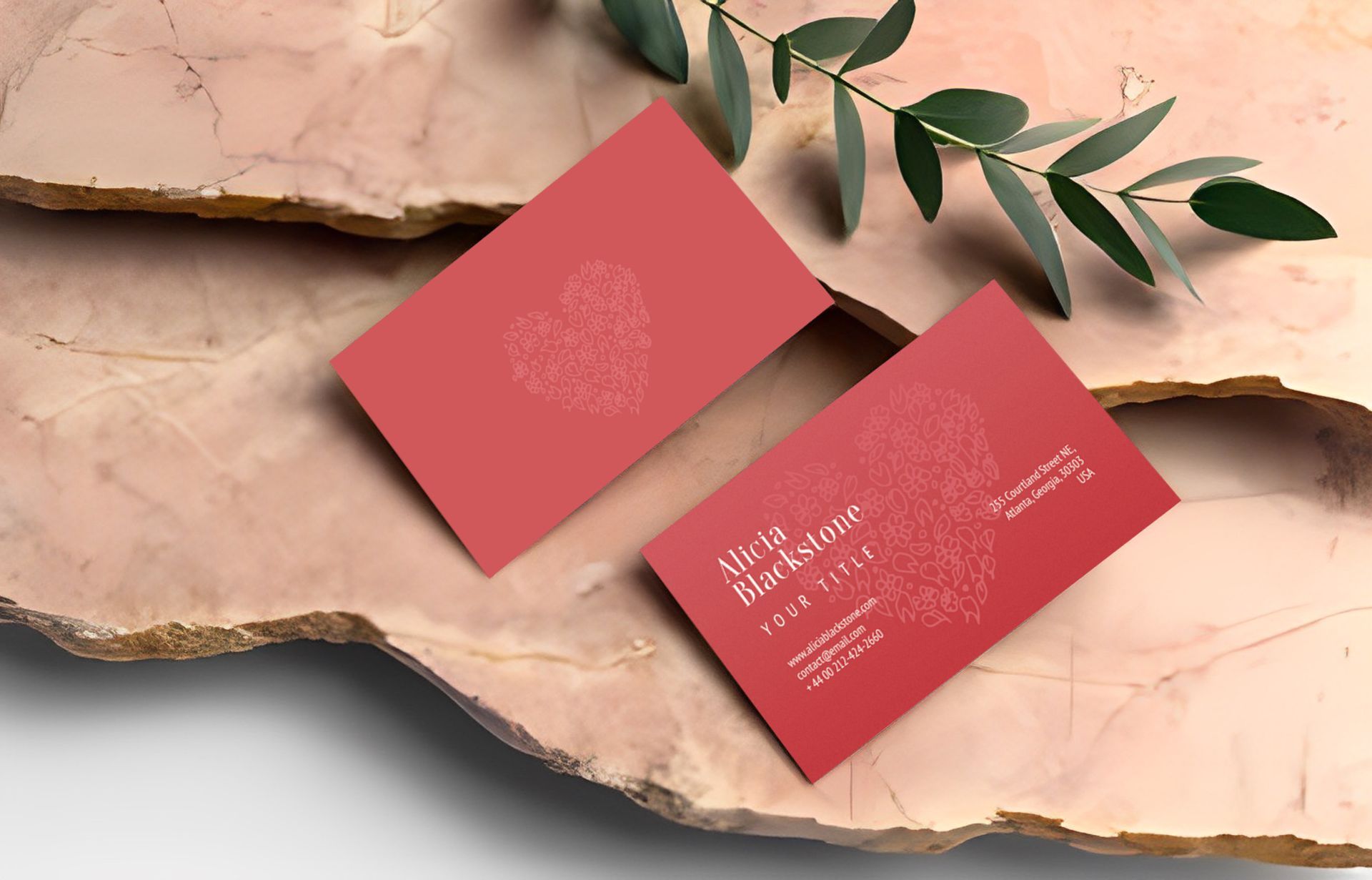 Two red business cards on a pink stone with a sprig of leaves. One card has text, both have a heart.