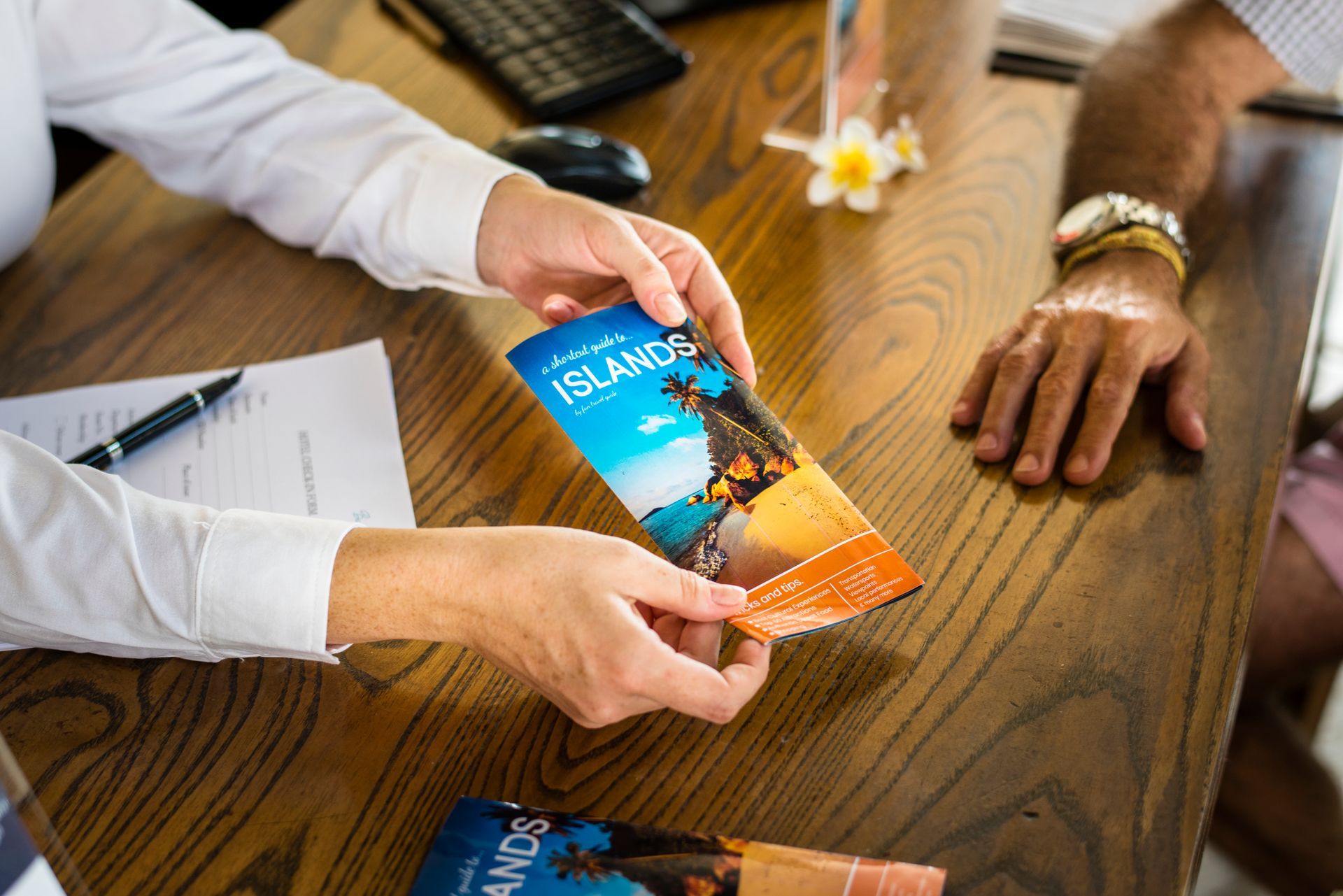 Hands holding a travel brochure with ocean scene at a wooden desk; another person is at right.