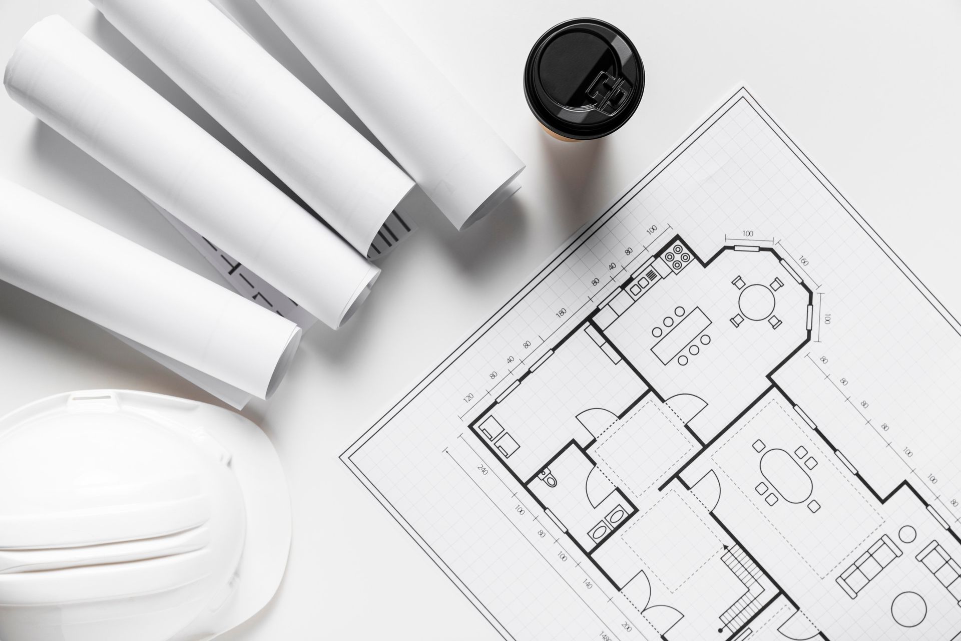 Architect's workspace with rolled blueprints, a floor plan, hard hat, and coffee cup on a white surface.