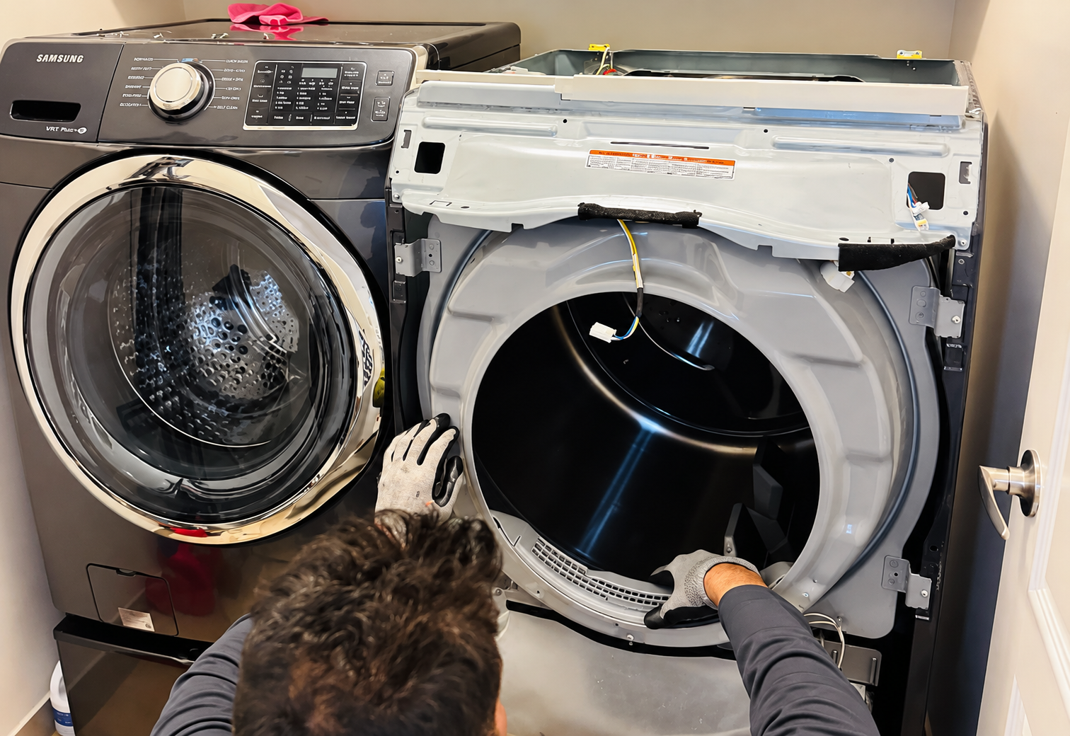 Two front-load washers being repaired, one open with a person’s hand inside and a cat watching nearby