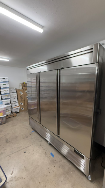 Large stainless-steel commercial refrigerator in a warehouse room with fluorescent lighting