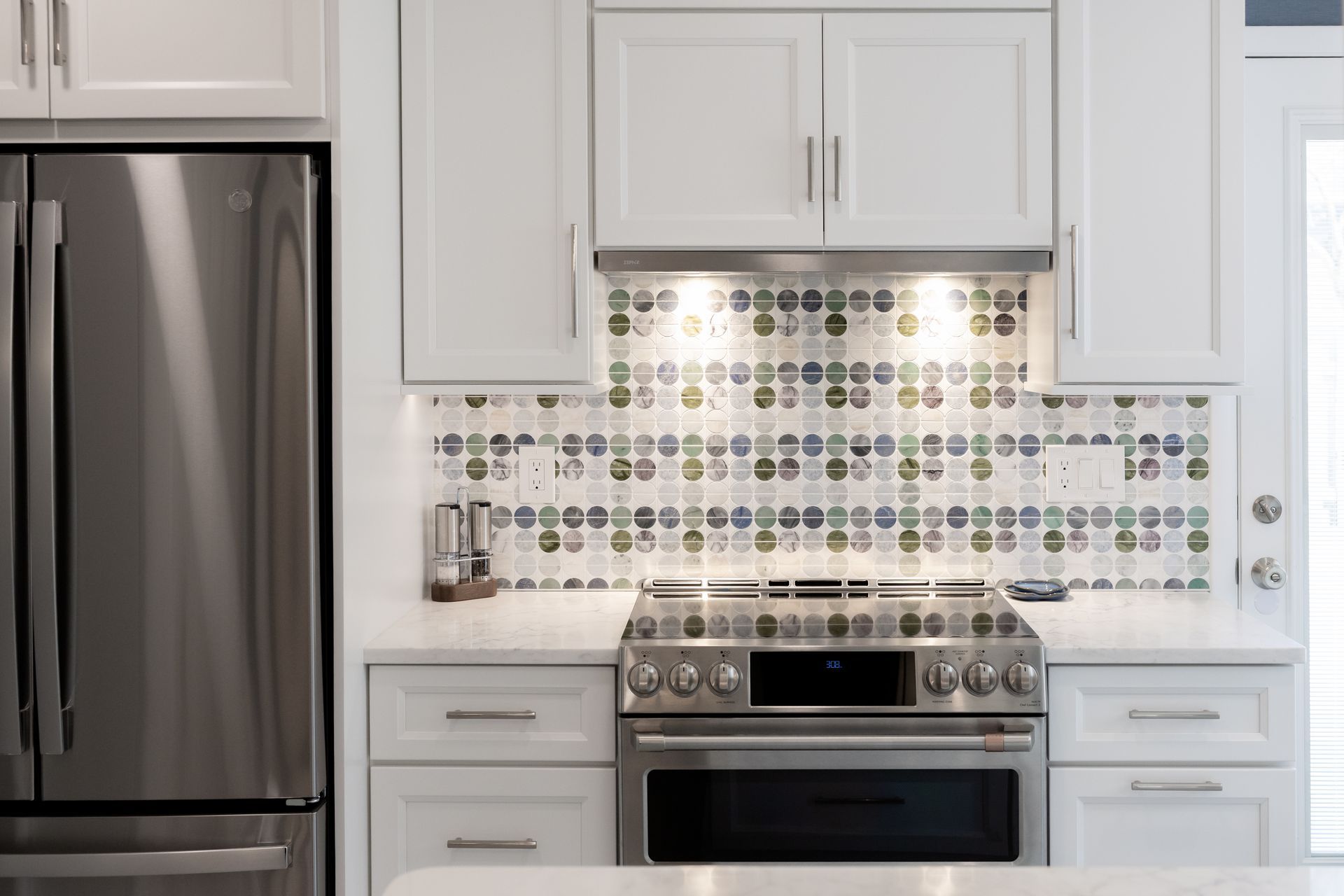 A kitchen with white cabinets and a stainless steel refrigerator and stove with designer tile behind stove.