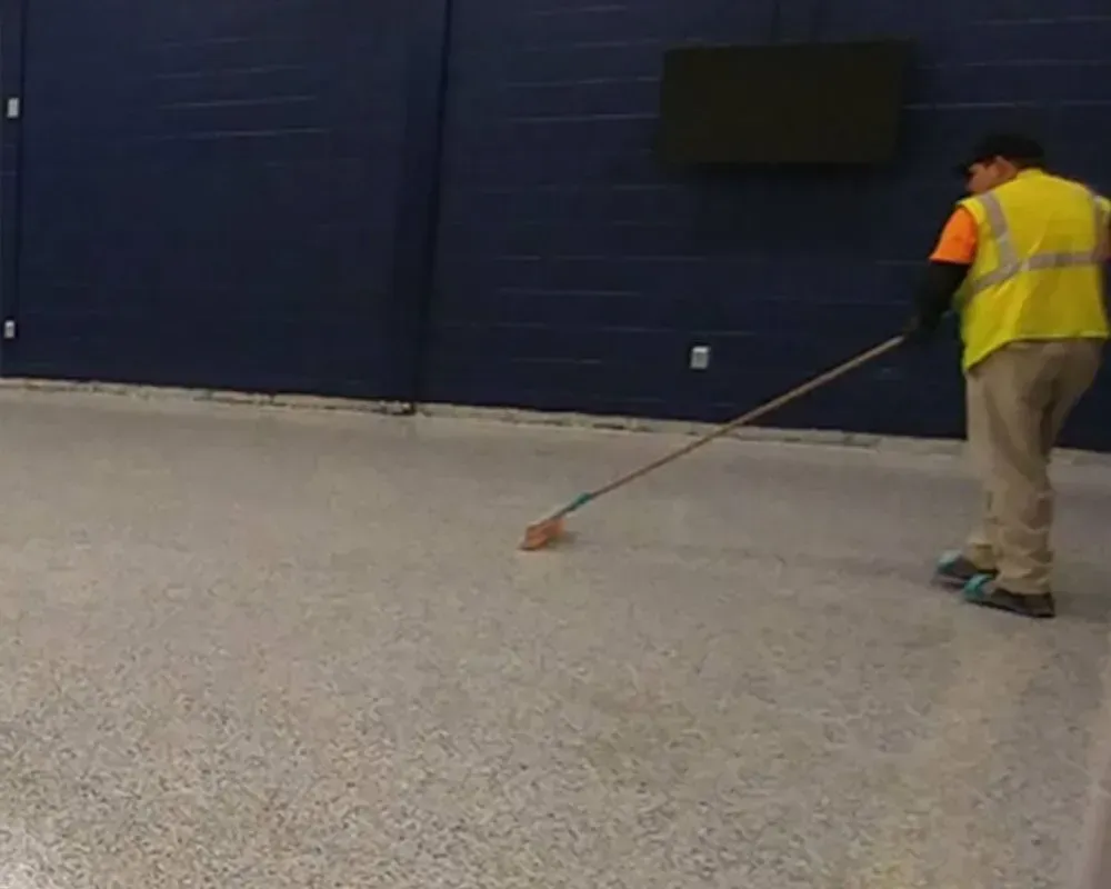 Worker in safety vest cleaning speckled floor with a long handled squeegee in a large room with dark blue wall.