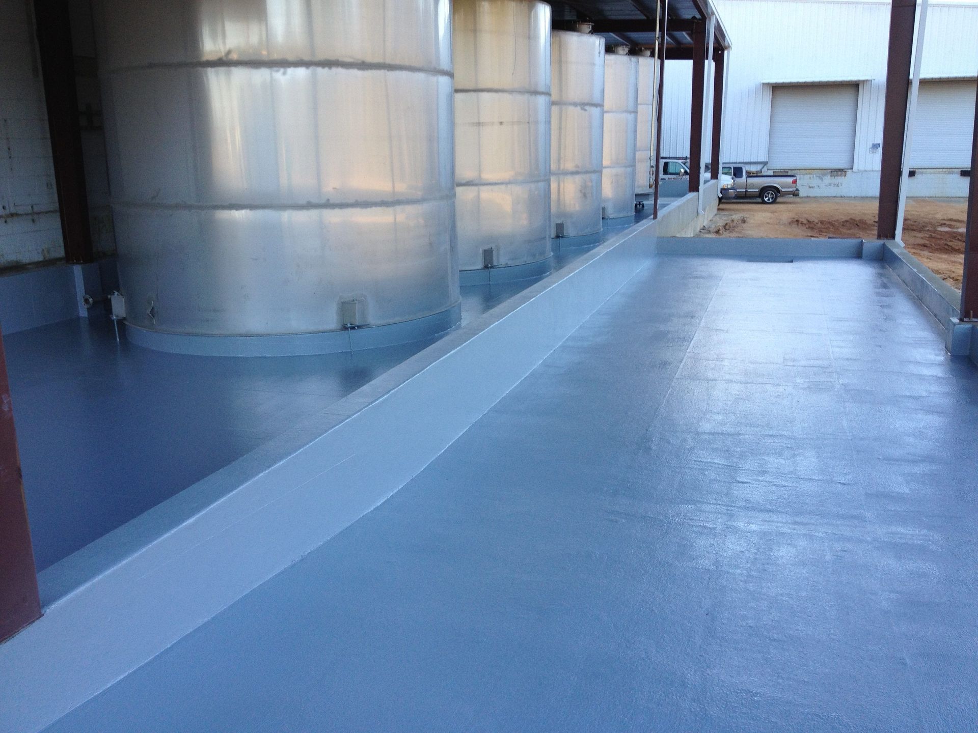 Metallic tanks on a grey, sealed concrete floor with a raised containment edge. Exterior setting.