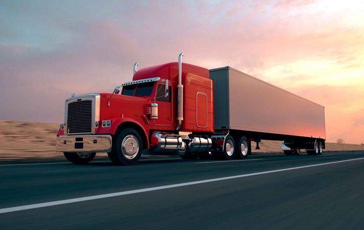 A red semi truck is driving down a highway at sunset.