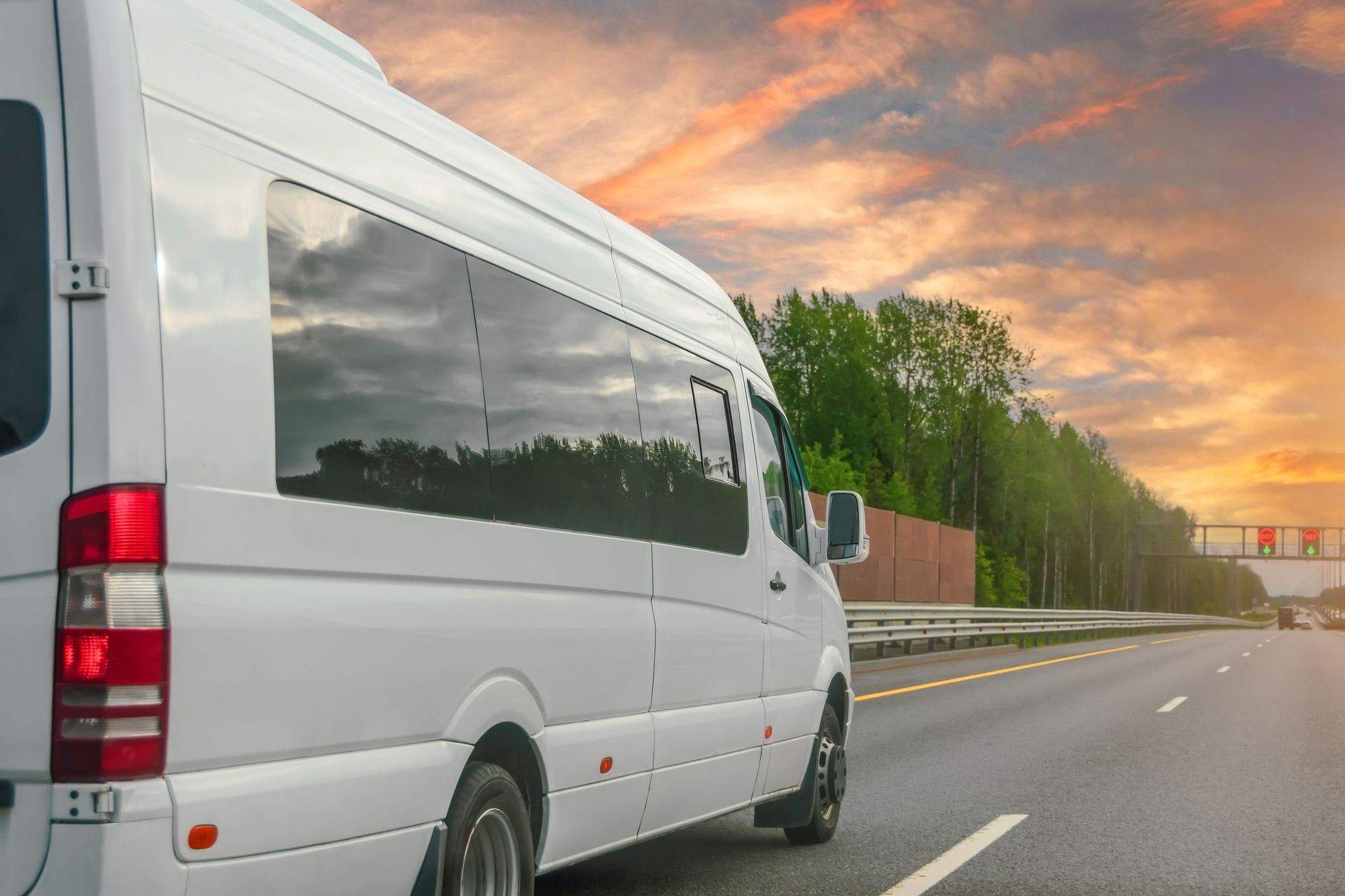 White passenger van driving on a road at sunset beside trees and a fence