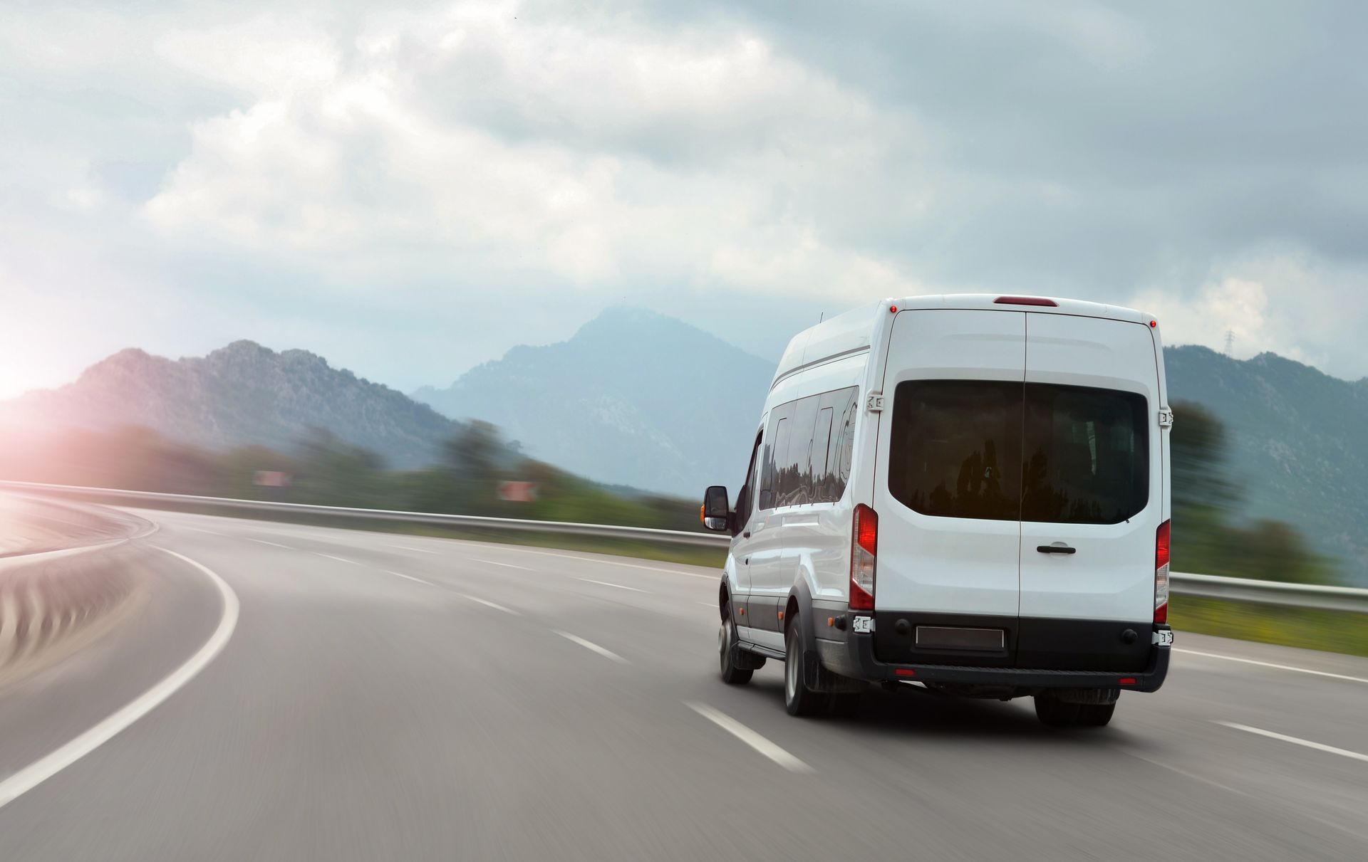White van driving on a curving highway with mountains in the background at sunrise.