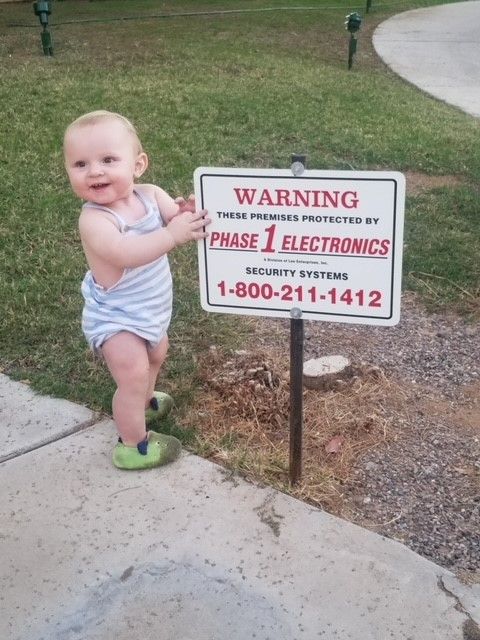 Smiling baby in a striped romper stands by a warning sign on a grassy sidewalk.