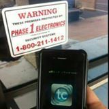 Smartphone in front of a red-and-white warning sign for Phase 1 Electronics with a phone number.