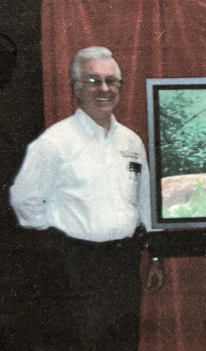 Smiling man in a white shirt stands beside a framed landscape painting